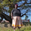 Activist Duduzile Dludla of Makhasaneni, standing in front of her home and an umbombe or forest strangler-fig (Ficus natalensis)